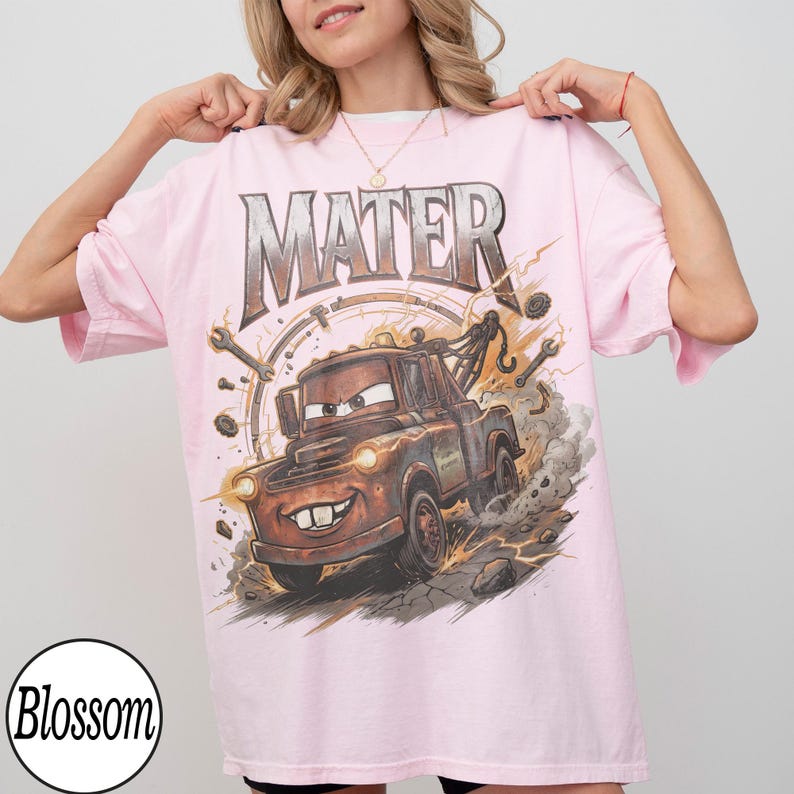 May include: Light pink t-shirt featuring a graphic of Mater from the Cars movie. The word "MATER" is printed above the cartoon tow truck. The design includes tools and lightning bolts, with the "Blossom" logo in the bottom left corner.