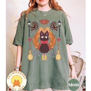 May include: Moss green t-shirt featuring a cartoon cat wearing a red bow tie, surrounded by decorative elements like a wreath, brooms, and street lamps. The shirt has the text "VELMIRASHOP SINCE 2025" and "Moss".