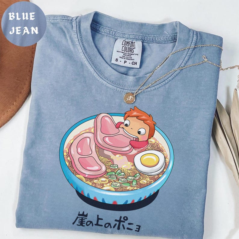 Puede incluir: Camiseta azul vaquero Comfort Colors con una ilustraci&oacute;n de dibujos animados de un personaje en un cuenco de ramen con cerdo, huevo y cebolletas. El texto japon&eacute;s "崖の上のポニョ" est&aacute; impreso en la parte inferior del cuenco. Un collar dorado est&aacute; sobre la camiseta.
