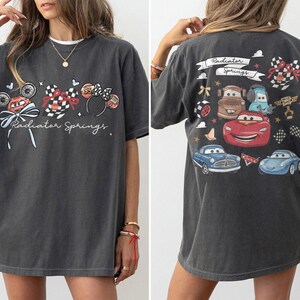 May include: Dark gray t-shirt featuring a front graphic of a race car, Mickey Mouse ears, and the text "Radiator Springs." The back of the shirt displays various cartoon cars and the words "Radiator Springs."