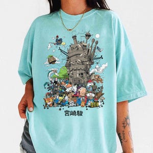 以下が含まれることがあります： 水色のTシャツ。カラフルな城と様々なキャラクターのイラストが描かれています。グラフィックには、下部に日本語の「宮崎駿」の文字が含まれています。柔らかい素材で作られたシャツです。