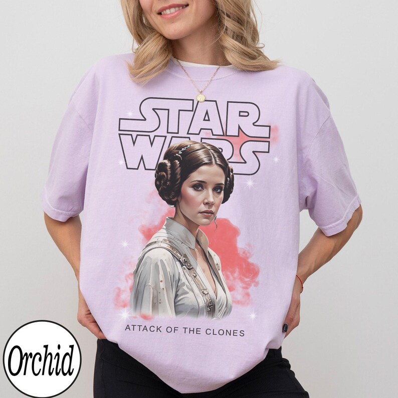 May include: A light purple t-shirt featuring a graphic of Princess Leia from Star Wars. The Star Wars logo is above the image of Leia, with the text "ATTACK OF THE CLONES" below. The shirt is a relaxed fit.