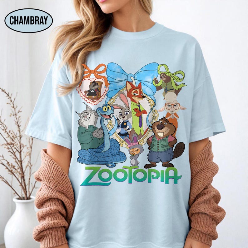 Puede incluir: Camiseta azul claro con un gr&aacute;fico colorido de personajes de la pel&iacute;cula animada Zootopia. El dise&ntilde;o incluye un lazo azul, un marco en forma de coraz&oacute;n y la palabra "Zootopia" en verde y azul. La camiseta tiene cuello redondo y mangas cortas.