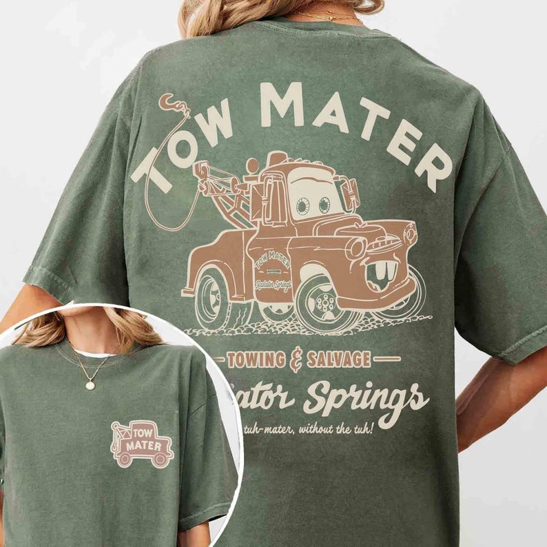 May include: A sage green t-shirt featuring a graphic of a brown tow truck on the back and a smaller matching graphic on the front left chest. The back design includes the text Tow Mater, Towing & Salvage, Radiator Springs, and tuh-mater, without the tuh.