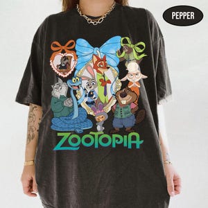 Puede incluir: Camiseta gris oscuro con un gr&aacute;fico colorido de personajes de la pel&iacute;cula animada Zootopia. El dise&ntilde;o incluye varios personajes de animales, un gran lazo azul y la palabra "Zootopia" en verde azulado y verde.