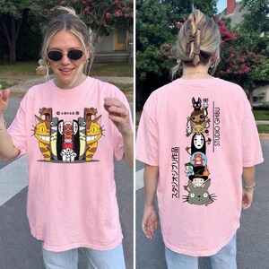 May include: Pink t-shirt featuring a colorful graphic of animated characters. The front design includes smiling cat-like creatures and other figures. The back of the shirt displays a vertical stack of characters with the text "STUDIO GHIBLI" and Japanese text.
