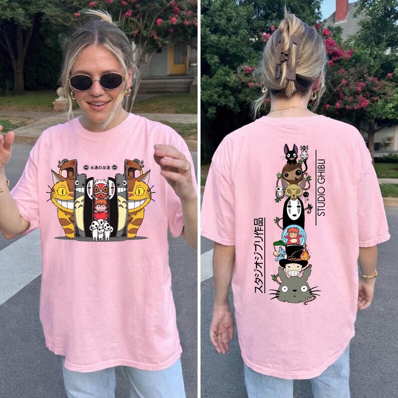 May include: Pink t-shirt featuring a colorful graphic of animated characters. The front design includes smiling cat-like creatures and other figures. The back of the shirt displays a vertical stack of characters with the text "STUDIO GHIBLI" and Japanese text.