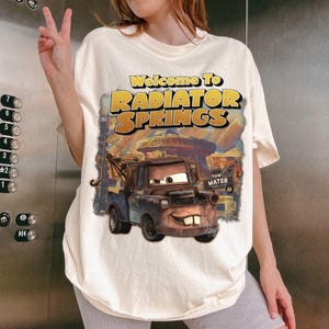 Puede incluir: Camiseta color crema con un gr&aacute;fico de Mate de la pel&iacute;cula Cars, con el texto "Welcome to Radiator Springs". El dise&ntilde;o incluye una ilustraci&oacute;n detallada del personaje y el nombre de la ciudad.