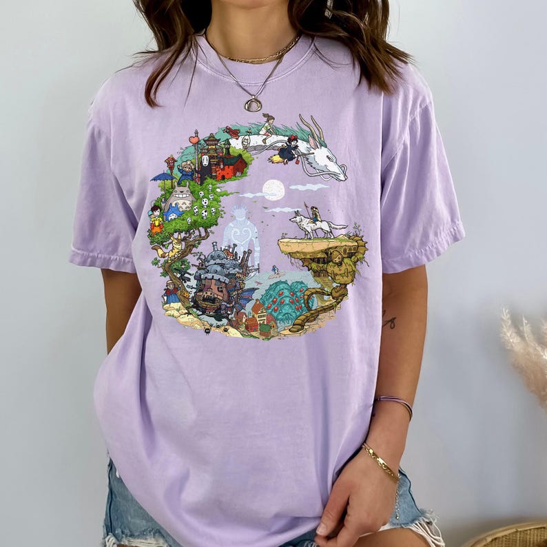 May include: A light purple t-shirt featuring a colorful graphic design. The design includes various cartoon characters and elements, such as a dragon, a castle, and other fantasy-themed illustrations. The shirt has short sleeves and a relaxed fit.