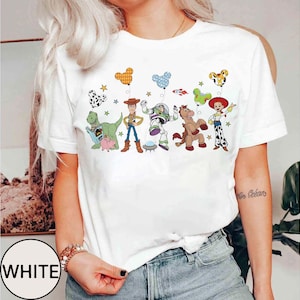 Puede incluir: Camiseta blanca con un gr&aacute;fico colorido de personajes de Toy Story, incluyendo a Woody y Buzz Lightyear. El dise&ntilde;o incluye globos y estrellas. La palabra "WHITE" est&aacute; impresa en un c&iacute;rculo blanco en la parte inferior.