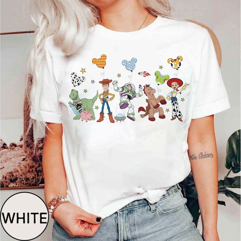 Puede incluir: Camiseta blanca con un gr&aacute;fico colorido de personajes de Toy Story, incluyendo a Woody y Buzz Lightyear. El dise&ntilde;o incluye globos y estrellas. La palabra "WHITE" est&aacute; impresa en un c&iacute;rculo blanco en la parte inferior.
