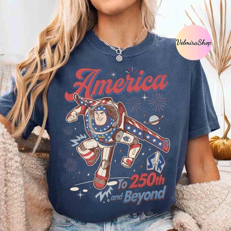 May include: Navy blue t-shirt with a graphic of a cartoon astronaut in a patriotic design. The text "America" is above the astronaut, and "To 250th and Beyond" is below. The design includes stars, fireworks, and a planet.