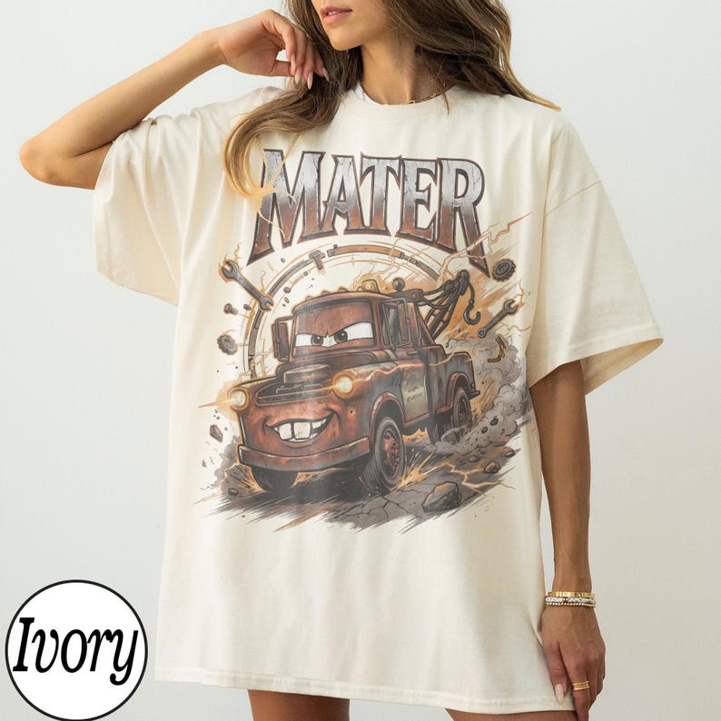 May include: Ivory-colored t-shirt featuring a graphic of Mater from the movie Cars. The design includes the word "MATER" above the truck, with a background of tools and a speedometer. The shirt is oversized.