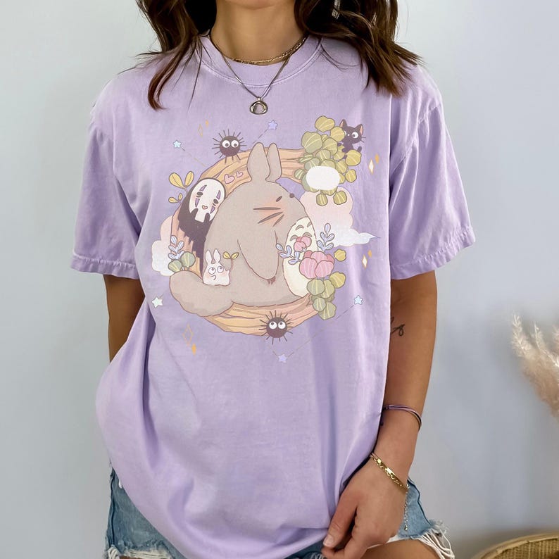 May include: A light purple t-shirt featuring a cartoon illustration of a large, gray, rabbit-like creature surrounded by smaller characters and floral elements. The design is set against a backdrop of stars and clouds.