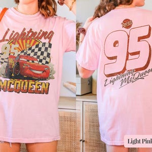 May include: A light pink t-shirt featuring a graphic of a red race car, a checkered flag, and the text Lightning McQueen on the front. The back of the shirt displays the number 95 in large font with the text Lightning McQueen below it.
