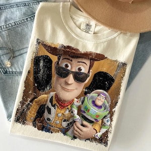Op de afbeelding: Een cr&egrave;mekleurig t-shirt met een grafische print van de personages Woody en Buzz Lightyear. Woody draagt een zwarte zonnebril en een bruine cowboyhoed. De afbeelding heeft een verweerde, textuurrijke uitstraling met een donkere rand.