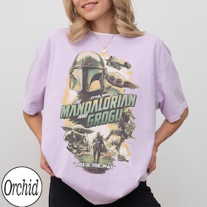 May include: A light purple t-shirt featuring a Star Wars graphic. The design includes the text "The Mandalorian and Grogu" with images of the Mandalorian, Grogu, and other characters. The phrase "This is the Way" is also printed on the shirt.