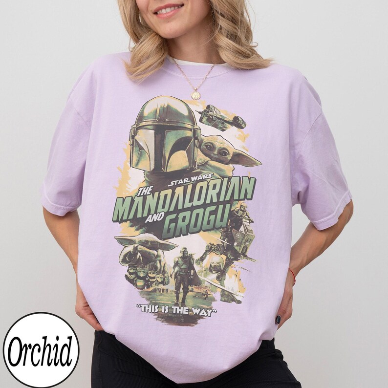 May include: A light purple t-shirt featuring a Star Wars graphic. The design includes the text "The Mandalorian and Grogu" with images of the Mandalorian, Grogu, and other characters. The phrase "This is the Way" is also printed on the shirt.