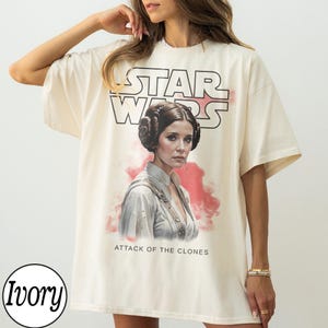 May include: Ivory-colored t-shirt featuring a graphic of Princess Leia from Star Wars, with the text "STAR WARS" above her portrait and "ATTACK OF THE CLONES" below. The shirt has short sleeves and a relaxed fit.