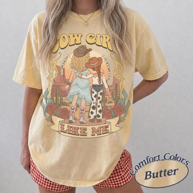 May include: A pale yellow short-sleeve t-shirt featuring a graphic of two figures in cowboy hats and boots standing in a desert landscape with cacti and roses. The text reads COW GIRL LIKE ME and I KNEW YOU I KNEW YOU. The shirt has a vintage wash.