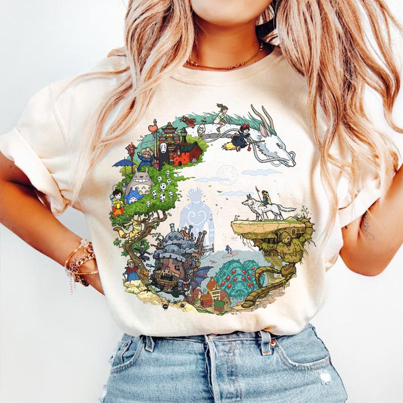 May include: Cream-colored t-shirt featuring a colorful circular graphic with animated characters and scenes. The design includes a white dragon, a castle, and various figures in a whimsical setting. The shirt is made of a soft material.