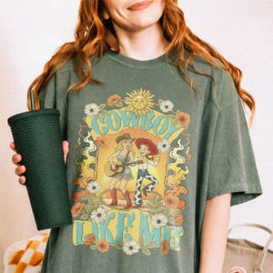 May include: A dark olive green short-sleeve t-shirt featuring a graphic print of two cartoon characters, one playing a guitar, surrounded by flowers and the text "COWBOY LIKE ME". A dark green studded tumbler with a straw is held in one hand.