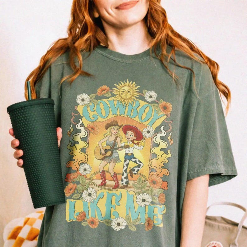May include: A dark olive green short-sleeve t-shirt featuring a graphic print of two cartoon characters, one playing a guitar, surrounded by flowers and the text "COWBOY LIKE ME". A dark green studded tumbler with a straw is held in one hand.