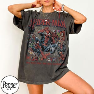 May include: Dark gray Spider-Man graphic t-shirt. The shirt features a collage of Spider-Man illustrations, the words "SPIDER-MAN" in red, and a spider web design. The shirt is a casual, oversized fit.