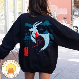 May include: Black sweatshirt featuring a stylized illustration of a white and blue goat-like creature with a red accent, set against a mountain and cloud backdrop. Japanese text is visible on the lower left. The sweatshirt has long sleeves.
