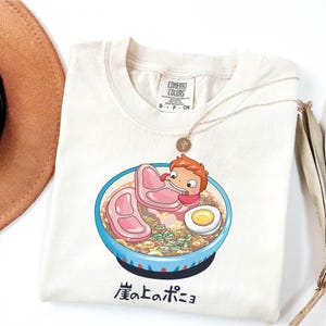 Puede incluir: Camiseta color crema con un cuenco de ramen de dibujos animados con un personaje de la pel&iacute;cula de animaci&oacute;n Ponyo. El cuenco es azul con un borde blanco y negro, y el ramen incluye carne rosa, un huevo y cebolletas. Un collar dorado descansa sobre la camiseta.