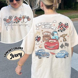 May include: Ivory-colored t-shirt featuring a graphic of characters and elements from the movie "Cars." The design includes the text "Radiator Springs" and various car illustrations. The shirt also has a "Comfort Colors" label.