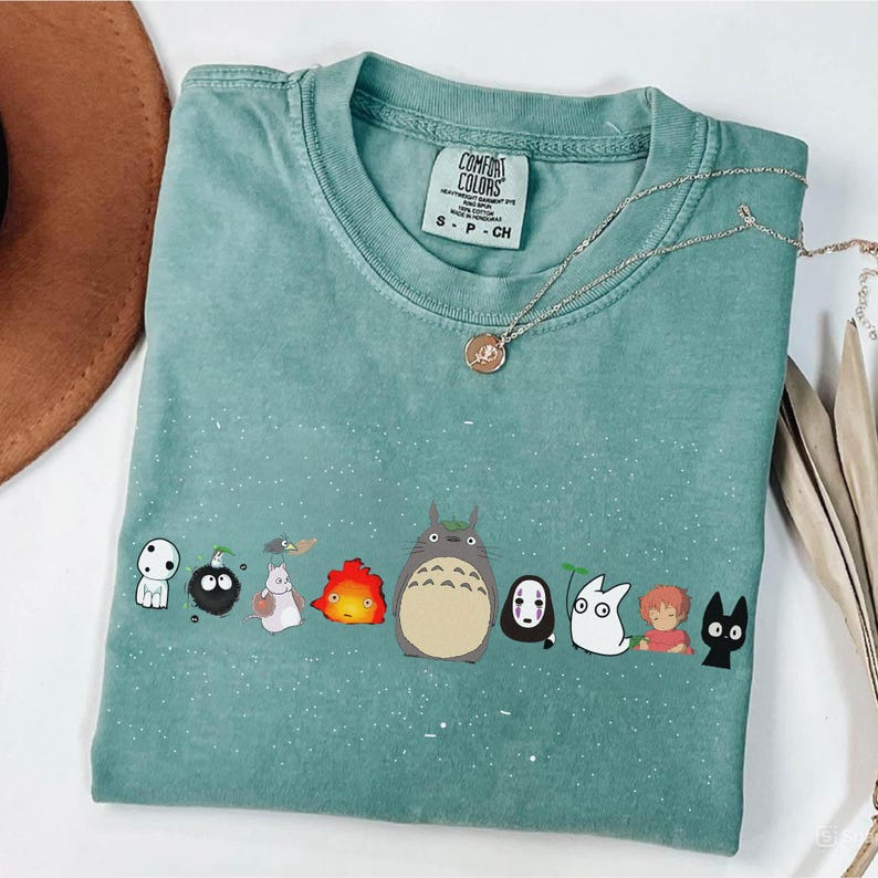 May include: A seafoam green Comfort Colors t-shirt featuring a line of cartoon characters. A gold necklace with a pendant rests on the shirt. The shirt has a small tag at the neckline with the brand name and size.