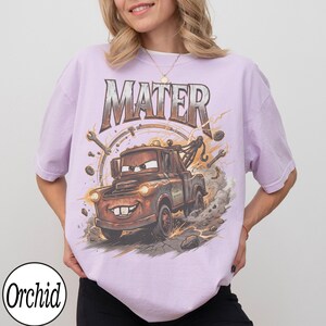 May include: A light purple t-shirt featuring a cartoon image of Mater from the movie Cars. The graphic includes the word "MATER" in a distressed font, along with wrenches and a speedometer design. The tow truck is brown with a smiling face.