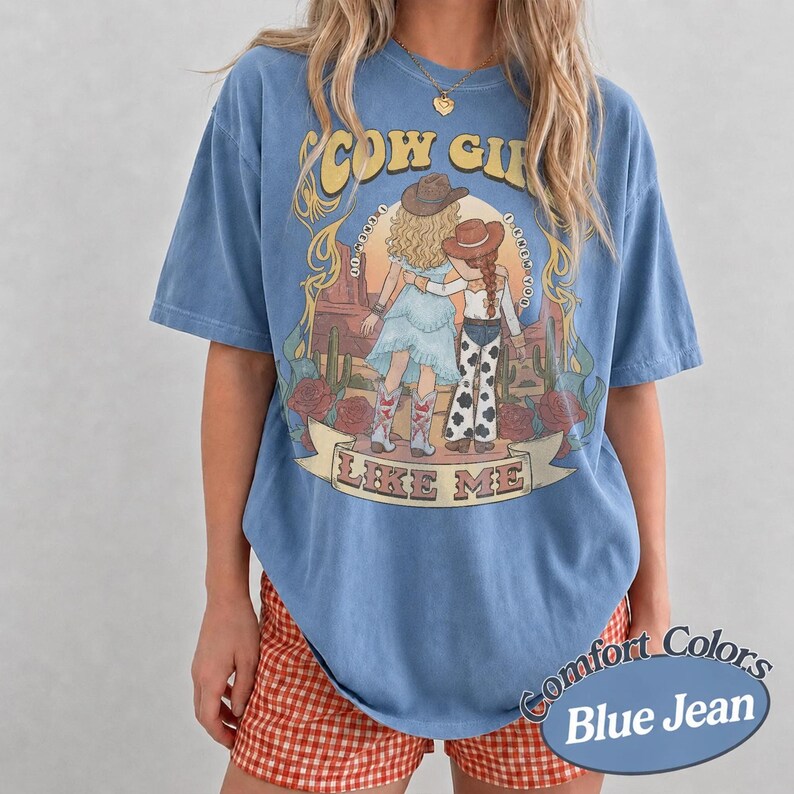 May include: A blue short-sleeve t-shirt featuring a graphic print of two figures in cowboy hats standing in a desert landscape. The text "COW GIRL" and "LIKE ME" is printed in yellow and brown. The shirt has a crew neckline and loose fit.