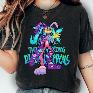 Könnte beinhalten: Ein anthrazitfarbenes T-Shirt mit einer Grafik eines lila und pinkfarbenen, hasenähnlichen Charakters mit mehreren Augen und Galaxienmuster. Der Text THE AMAZING DIGITAL CIRCUS steht in leuchtend blauen und grünen Buchstaben vor einem bunten Hintergrund.