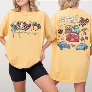 May include: Yellow t-shirt featuring a front graphic of Mickey Mouse ears, checkered patterns, and the text "Radiator Springs." The back of the shirt displays various car illustrations and the words "Radiator Springs" in a banner.