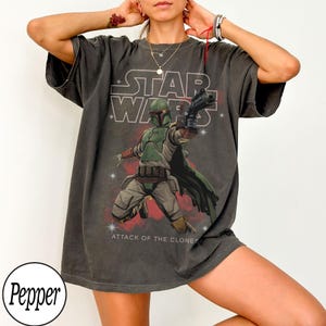 May include: Dark gray t-shirt featuring a Star Wars graphic. The design includes the Star Wars logo, an illustration of Boba Fett, and the text "Attack of the Clones". The shirt has short sleeves and a relaxed fit.