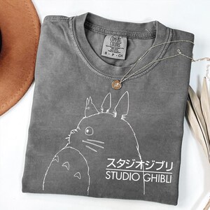 May include: A gray Comfort Colors t-shirt featuring a white graphic of a Studio Ghibli character and the text "STUDIO GHIBLI" in English and Japanese. A silver necklace with a pendant and a brown hat are also visible.