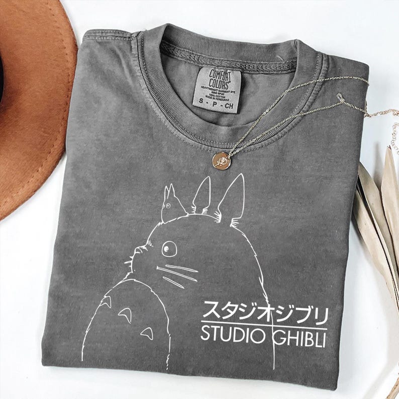 May include: A gray Comfort Colors t-shirt featuring a white graphic of a Studio Ghibli character and the text "STUDIO GHIBLI" in English and Japanese. A silver necklace with a pendant and a brown hat are also visible.