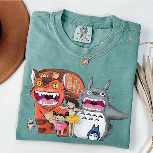 May include: A seafoam green Comfort Colors t-shirt featuring a graphic of characters from the animated film "My Neighbor Totoro." The design includes the Catbus, Totoro, and two young girls. A gold necklace and a brown hat are also visible.