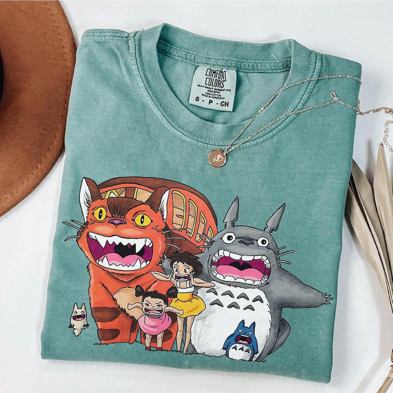 May include: A seafoam green Comfort Colors t-shirt featuring a graphic of characters from the animated film "My Neighbor Totoro." The design includes the Catbus, Totoro, and two young girls. A gold necklace and a brown hat are also visible.