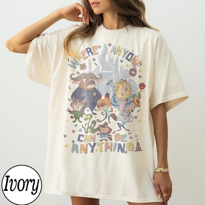 May include: Ivory-colored t-shirt featuring a colorful graphic of cartoon characters and the text "WHERE ANYONE CAN BE ANYTHING." The design includes a cityscape and various animal figures. The shirt has short sleeves and a relaxed fit.