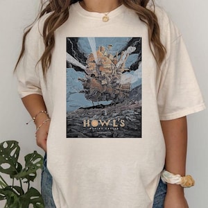 Puede incluir: Camiseta color crema con un gr&aacute;fico de la pel&iacute;cula de animaci&oacute;n "El castillo ambulante". El dise&ntilde;o muestra el castillo en tonos marrones y dorados sobre un cielo azul. El texto "HOWL'S MOVING CASTLE" est&aacute; en la parte inferior.