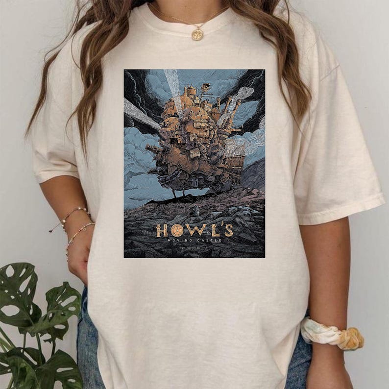Puede incluir: Camiseta color crema con un gr&aacute;fico de la pel&iacute;cula de animaci&oacute;n "El castillo ambulante". El dise&ntilde;o muestra el castillo en tonos marrones y dorados sobre un cielo azul. El texto "HOWL'S MOVING CASTLE" est&aacute; en la parte inferior.
