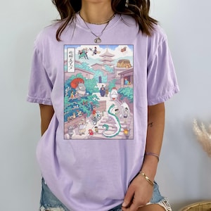 May include: A light purple t-shirt featuring a colorful anime-style illustration. The graphic depicts a street scene with various characters, buildings, and a dragon. The shirt is made of a soft material and has a relaxed fit.