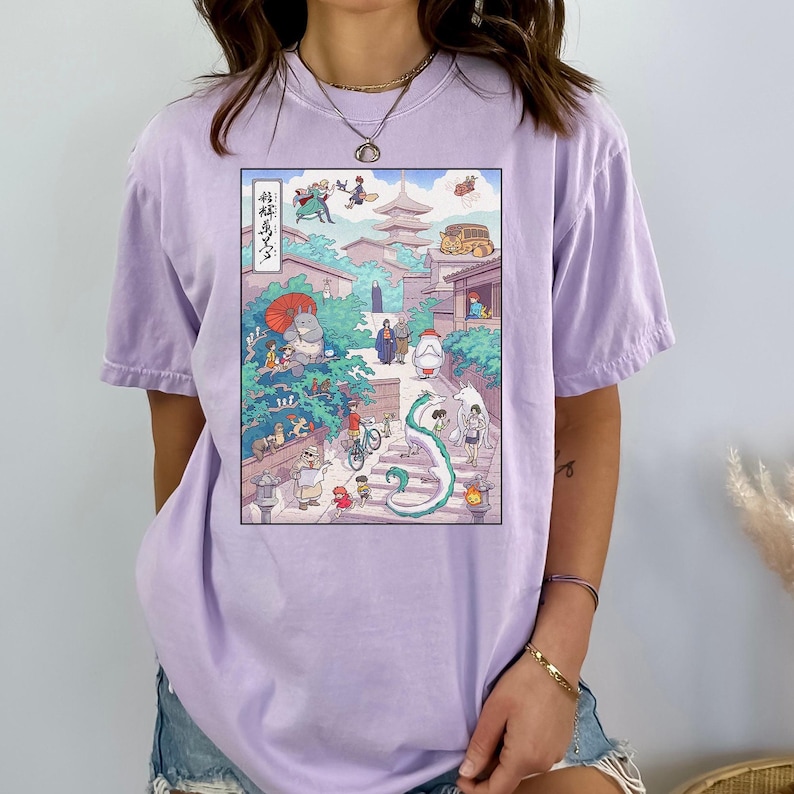 May include: A light purple t-shirt featuring a colorful anime-style illustration. The graphic depicts a street scene with various characters, buildings, and a dragon. The shirt is made of a soft material and has a relaxed fit.