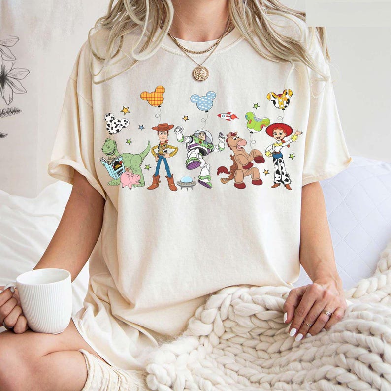 Puede incluir: Camiseta color crema con un gr&aacute;fico colorido de personajes animados y globos. El dise&ntilde;o incluye un dinosaurio, un vaquero, un astronauta, un caballo y una vaquera. La camiseta es de estilo casual y oversize.