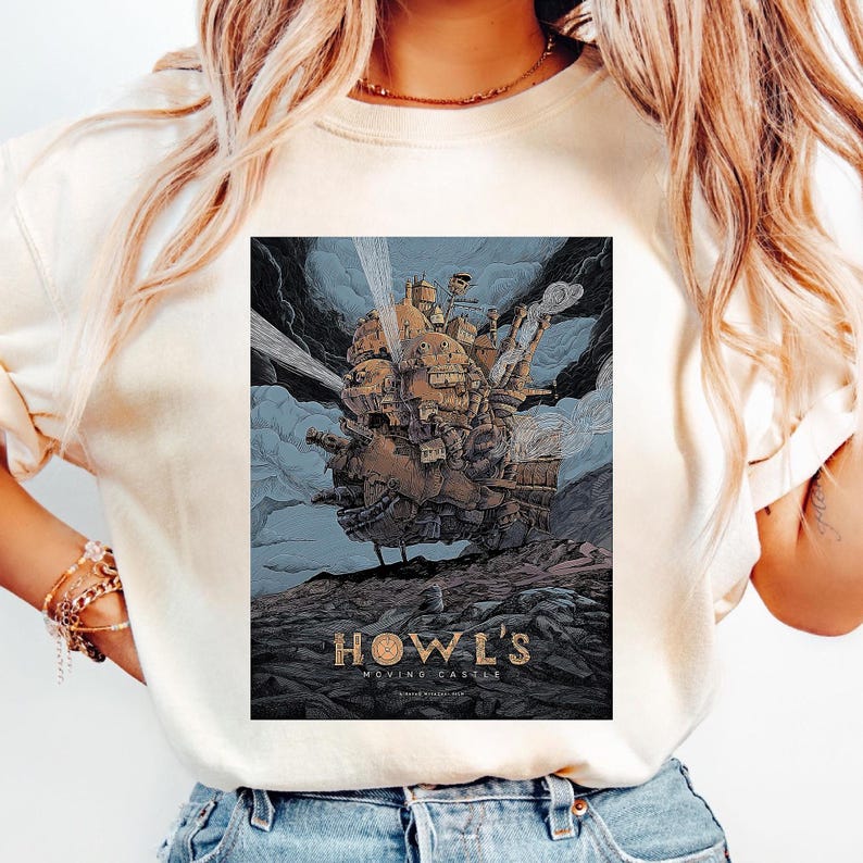 Puede incluir: Camiseta color crema con un dise&ntilde;o gr&aacute;fico de la pel&iacute;cula de animaci&oacute;n "El castillo ambulante". El dise&ntilde;o muestra el castillo con un cielo azul y nubes oscuras. El texto "HOWL'S MOVING CASTLE" est&aacute; impreso en dorado.