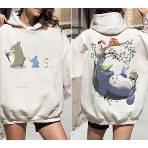 Op de afbeelding: Crèmekleurige hoodie met een grafische afbeelding van animatiefiguren op de voorkant, waaronder een groot grijs wezen met een paraplu. De achterkant van de hoodie toont een gedetailleerde illustratie van een boslandschap met extra figuren en een boom.