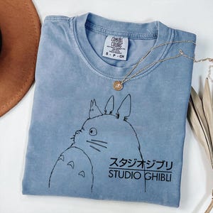 May include: A light blue t-shirt featuring a black graphic of a cartoon character and Japanese text. The text reads "STUDIO GHIBLI". A gold necklace with a pendant is draped across the shirt. The shirt is labeled "COMFORT COLORS".